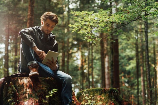 Young handsome man sitting in the woods and using a digital touch screen tablet, wi-fi connection and freedom concept